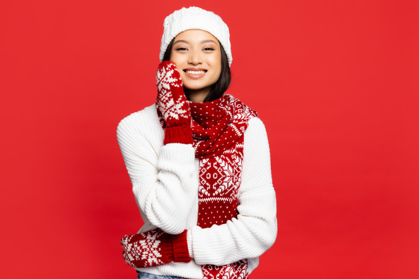 cheerful young asian woman in white hat, mittens and scarf smiling isolated on red  - Foto, Bild