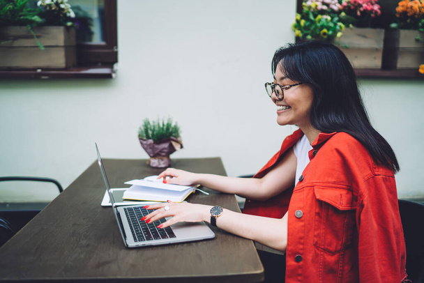 Side view of positive Asian female freelancer in casual outfit and eyewear sitting at wooden table and working on laptop - Φωτογραφία, εικόνα