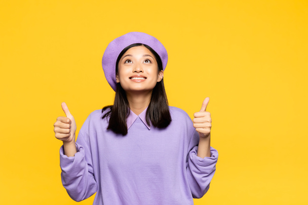 happy asian woman in beret showing thumbs up and looking up isolated on yellow - Photo, Image