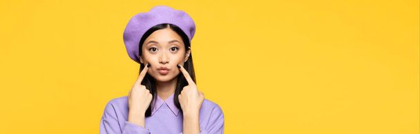 femme asiatique en béret toucher les joues avec les doigts tout en boudant les lèvres isolées sur jaune, bannière - Photo, image