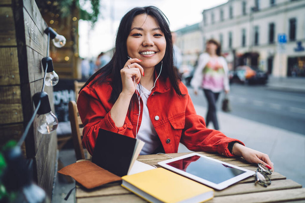 Cheerful Asian millennial female with earphones with built in microphone using tablet to communicate with friends while sitting in street cafe in summer and smiling at camera - Foto, afbeelding