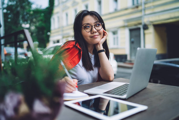 Young Asian female blogger in casual outfit and eyeglasses looking away dreamily while sitting at table with open laptop and tablet and taking notes in notepad - Φωτογραφία, εικόνα