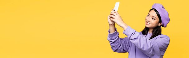 feliz asiático mujer en boina tomando selfie en smartphone aislado en amarillo, bandera - Foto, Imagen
