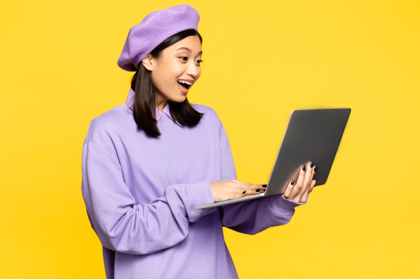 gelukkig aziatische vrouw in baret met behulp van laptop geïsoleerd op geel  - Foto, afbeelding