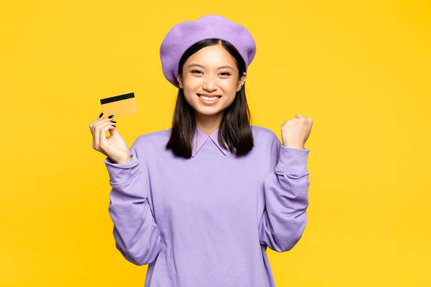 vzrušená asijská žena v baretu držící kreditní kartu a radující se izolované na žluté  - Fotografie, Obrázek