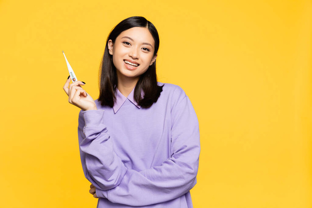 happy asian woman holding digital thermometer isolated on yellow  - Фото, изображение