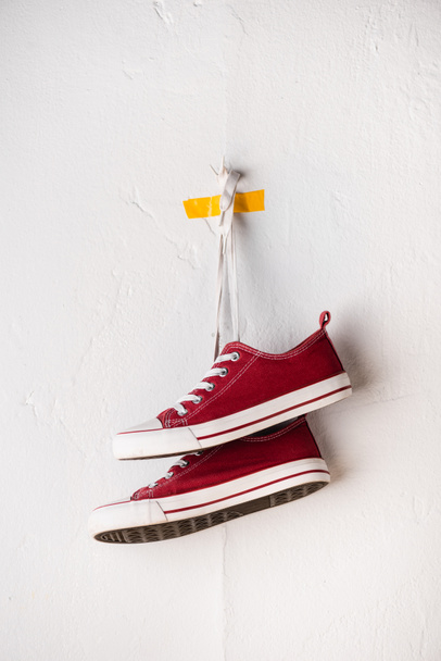 白いテクスチャの壁の近くの靴ひもにぶら下がっているカジュアルな赤いスニーカー - 写真・画像
