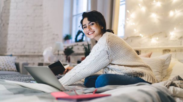 Full body optimistic woman in casual outfit sitting in lotus pose in bed and browsing laptop while working remotely at home - Zdjęcie, obraz