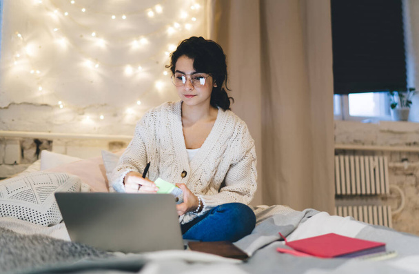 Positive female in glasses and pajama sitting on comfortable bed and browsing laptop while working on remote project at home - Foto, Imagen