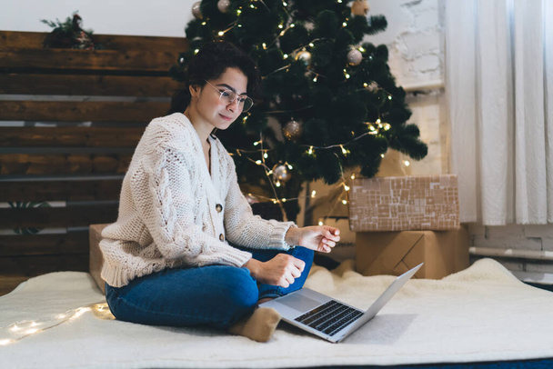 Full body of young lady in warm wear sitting with legs crossed on floor under Christmas tree and surfing laptop - Фото, зображення