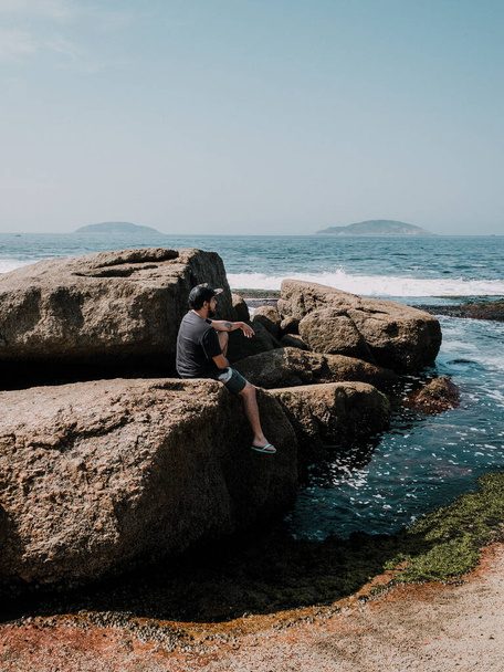 RIO DE JANEIRO, BRAZIL - Sep 10, 2020: Beautiful landscape shot of a guy sitting on a rock on the beach during a bright sky summer day. - Photo, image