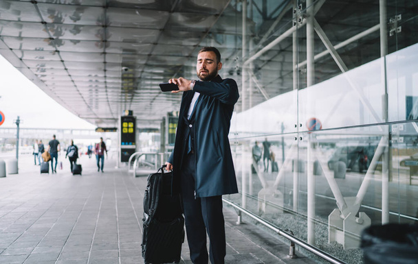 Bearded handsome businessman wearing formal with luggage standing near modern building and looking at clock waiting for transport during trip - Photo, Image