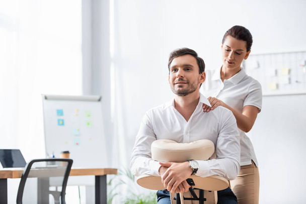 Massaggiatrice massaggiando spalla di uomo d'affari sorridente seduto su sedia da massaggio in ufficio su sfondo sfocato - Foto, immagini