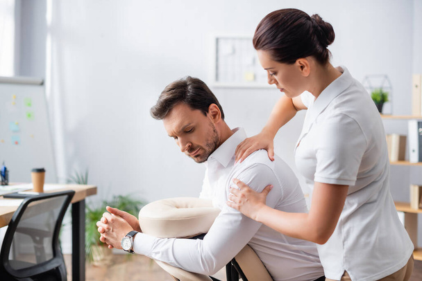 Female massage therapist massaging shoulder of client sitting on massage chair in office on blurred background - Fotografie, Obrázek