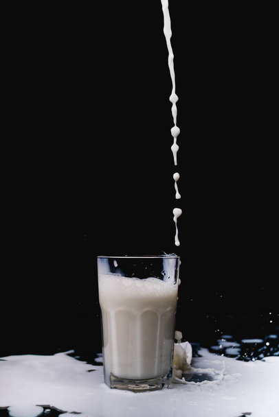 Ένα κοντινό πλάνο του γάλακτος που χύνεται σε ένα γυάλινο κύπελλο σε μαύρο φόντο - Φωτογραφία, εικόνα
