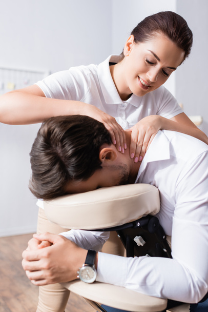 Sorridente massaggiatrice facendo seduto massaggio di uomo d'affari collo in ufficio su sfondo sfocato - Foto, immagini
