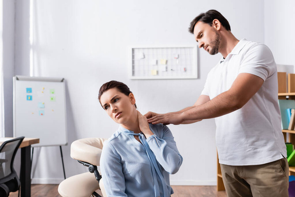 Masér stojí a dívá se na podnikatelku s bolestí v krku dívá pryč, zatímco sedí na masážním křesle v kanceláři - Fotografie, Obrázek