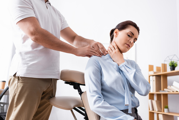 Massothérapeute massant l'épaule douloureuse de la femme d'affaires assise sur une chaise de massage au bureau sur fond flou - Photo, image