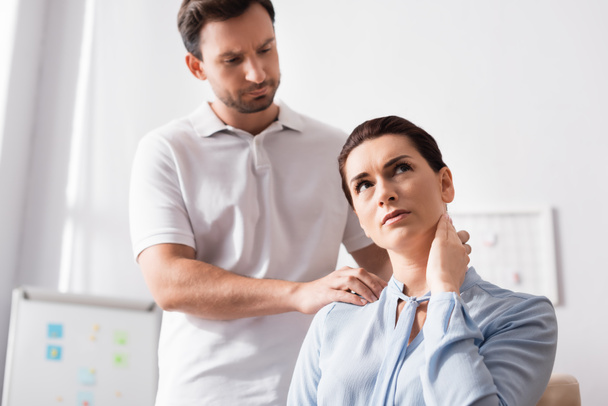 Focused masseur massaging hurting shoulder of female client with hand on neck in office on blurred background - Фото, зображення