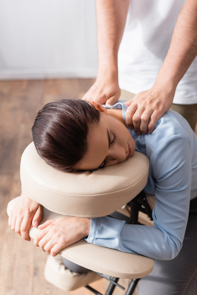Masér s nataženýma rukama masírující ramena brunetky podnikatelky sedící na masážním křesle - Fotografie, Obrázek