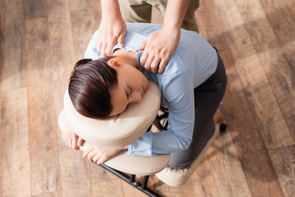 Pohled shora na masérku masírující ramena brunetky podnikatelky se zavřenýma očima, sedící na masážním křesle - Fotografie, Obrázek
