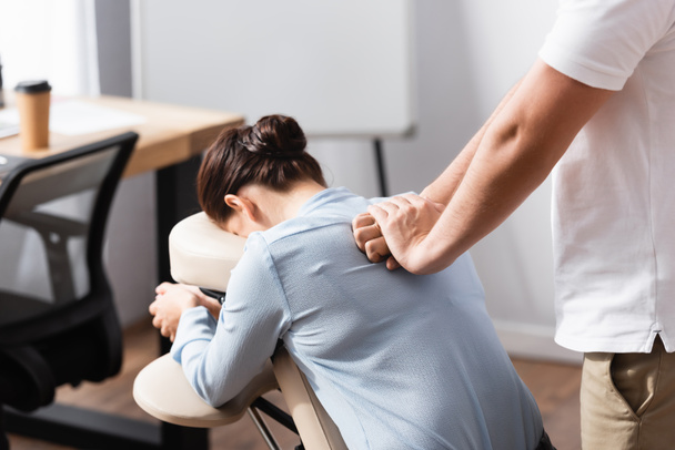 Massagista massageando mulher morena de volta sentado na cadeira de massagem com escritório borrado no fundo - Foto, Imagem