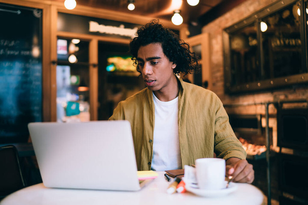 Focused ethnic man in casual clothes sitting at wooden table in modern cafe while working on remote project on laptop - Foto, imagen