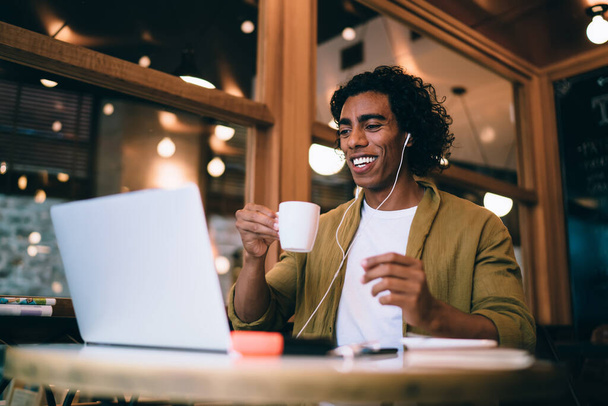 Prosperous handsome male freelancer discussing business ideas during online conversation using technology and earphones, smiling millennial businessman making video call in cafeteria - Photo, Image