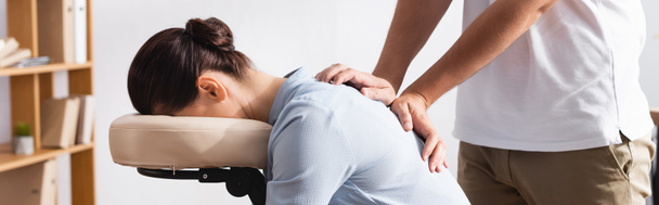 Side view of masseur massaging back of brunette woman sitting on massage chair on blurred background, banner - Photo, Image