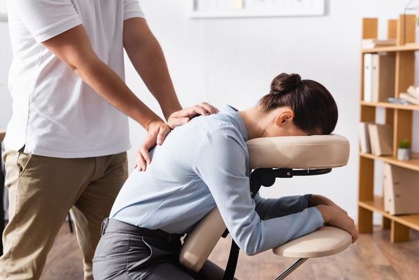 Side view of masseur doing seated massage of back for brunette businesswoman with blurred office on background - Fotografie, Obrázek