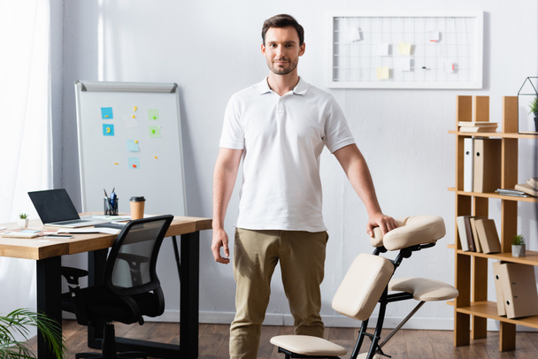 Front view of positive physiotherapist looking at camera while leaning on massage chair in office - Fotoğraf, Görsel