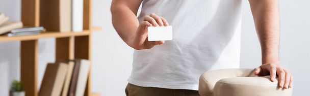 Cropped view of masseur showing blank business card, while standing near massage chair on blurred background, banner - Foto, immagini