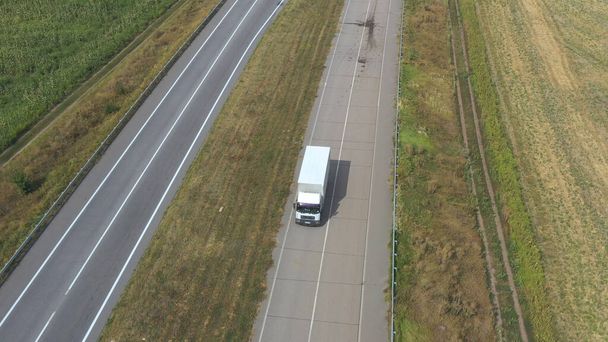 Aerial shot of truck with cargo trailer driving on empty road and transporting goods. Flying over delivery lorry moving along highway in countryside. Cargo transportation concept. Slow motion Top view. - Zdjęcie, obraz