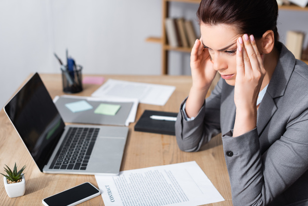 Businesswoman with migraine looking away while sitting near contracts and laptop on table on blurred background - Photo, Image