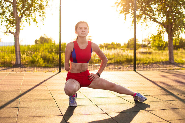 Young woman athlete with an amputated arm and burns on her body limber up on the sports field. Outdoor workout. - Photo, Image