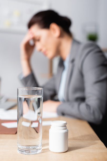 Nahaufnahme von Glas Wasser und Medikamenten auf dem Schreibtisch mit verschwommener Geschäftsfrau mit Kopfschmerzen im Hintergrund - Foto, Bild