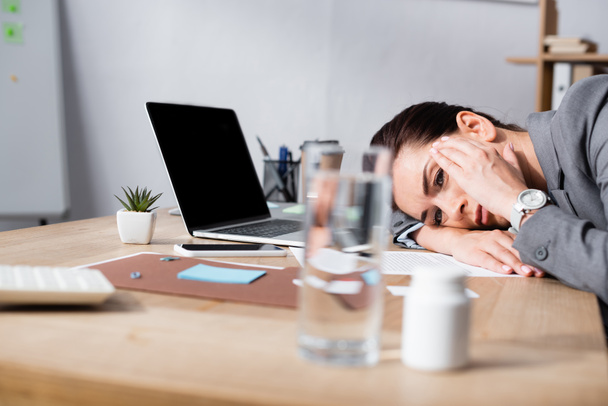 Деловая женщина с мигренью опирается на стол с ноутбуком в офисе на размытом переднем плане  - Фото, изображение