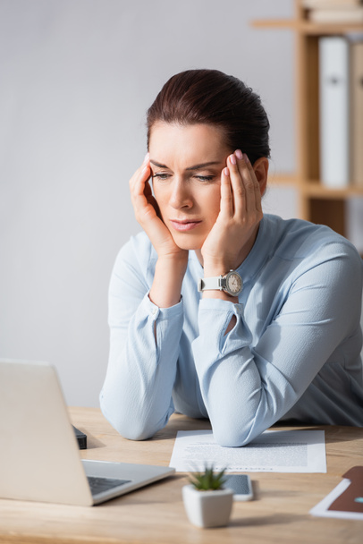 Müde brünette Geschäftsfrau mit Kopfschmerzen lehnt am Arbeitsplatz auf verschwommenem Hintergrund - Foto, Bild