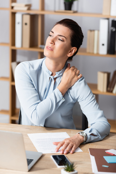 Κουρασμένη επιχειρηματίας με πόνο στον ώμο κοιτάζοντας μακριά, ενώ κάθεται στο χώρο εργασίας σε θολή φόντο - Φωτογραφία, εικόνα