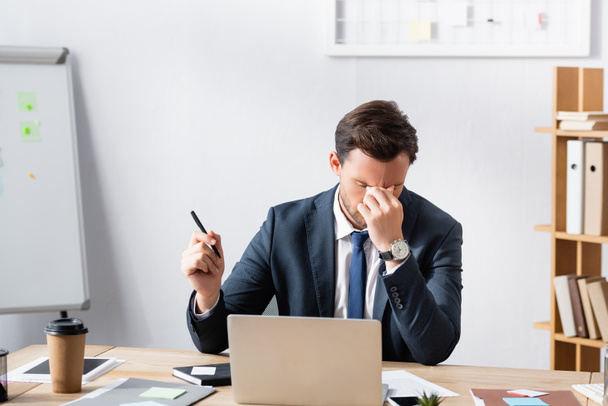 Hombre de negocios agotado con dolor de cabeza, cogido de la mano cerca de los ojos, sentado en el lugar de trabajo sobre fondo borroso - Foto, imagen