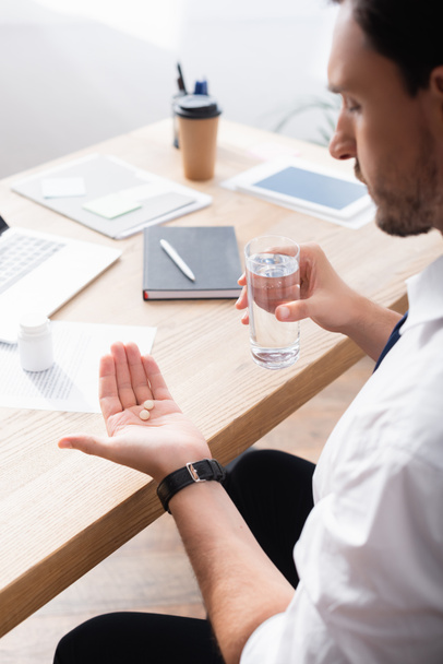 Empresario sosteniendo un vaso de agua y mirando píldoras en la palma de la mano, mientras está sentado en el lugar de trabajo sobre un fondo borroso - Foto, imagen