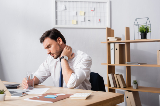 Üzletember fájdalmas nyakkal egy pohár vizet tartva, miközben a munkahelyén ül az irodában - Fotó, kép