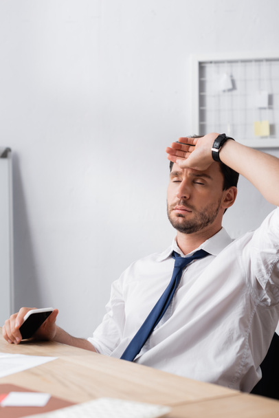 Exhausted businessman with closed eyes, holding hand on forehead, while sitting at workplace on blurred foreground - Foto, Imagem