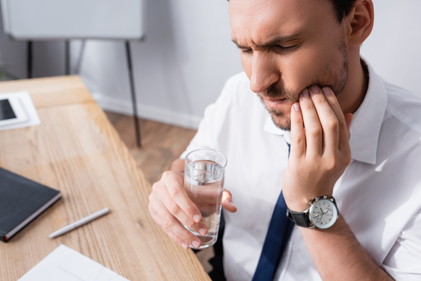 Geschäftsmann hält ein Glas Wasser in der Hand und leidet unter Zahnschmerzen, während er am Arbeitsplatz vor verschwommenem Hintergrund sitzt - Foto, Bild