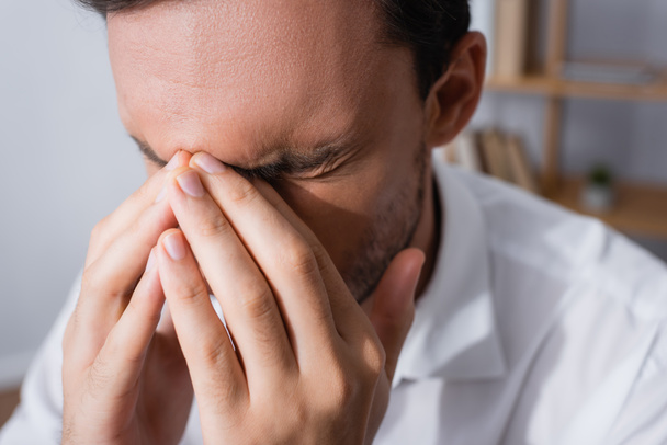 Close up widok wyczerpany pracownik biurowy z migreną, trzymając się za ręce w pobliżu zamkniętych oczu na niewyraźne tło - Zdjęcie, obraz