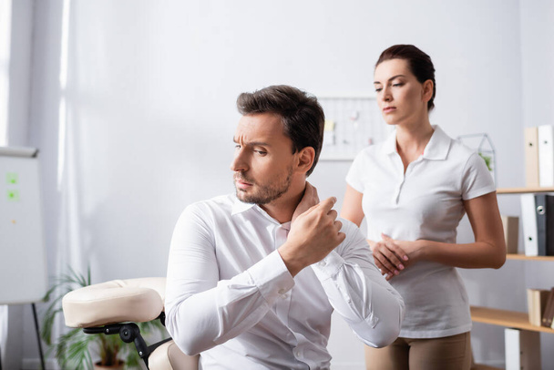 Masseuse looking at businessman pointing with finger at painful neck, while sitting on massage chair on blurred background - Photo, Image
