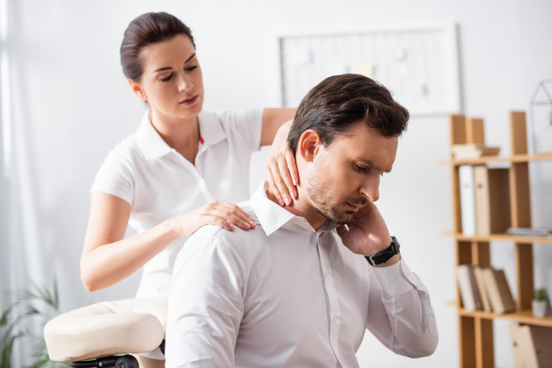 Massothérapeute féminine massant le cou douloureux de l'homme d'affaires assis sur une chaise de massage sur fond flou - Photo, image