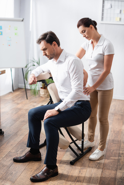 Massothérapeute féminine massant le dos douloureux de l'homme d'affaires assis sur une chaise de massage au bureau - Photo, image