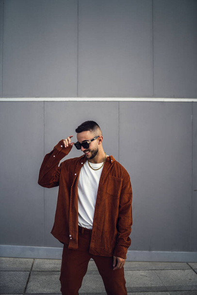 A vertical shot of a young handsome European man in a brown stylish outfit and sunglasses posing at the camera - Foto, afbeelding