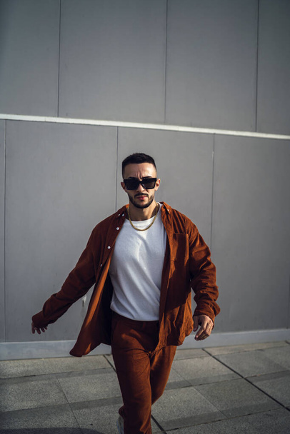A vertical shot of a young handsome European man in a brown stylish outfit and sunglasses posing at the camera - Фото, зображення